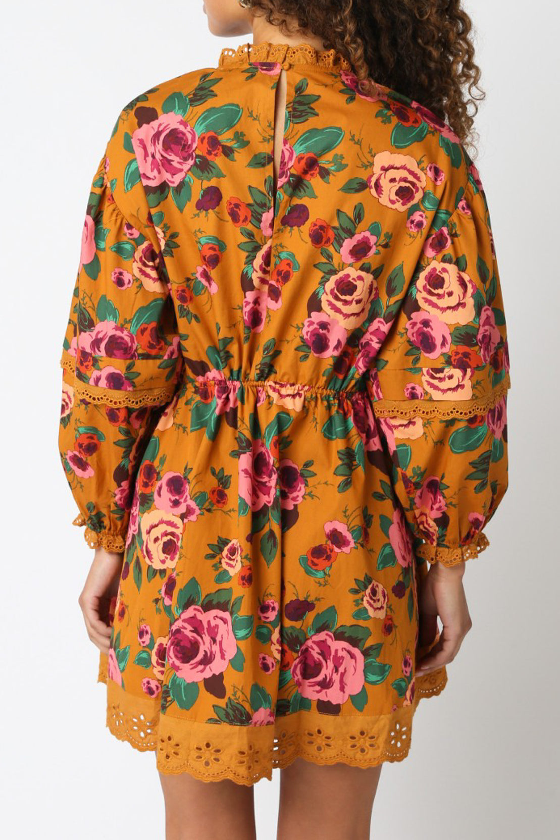 Back view of a woman wearing a mustard yellow floral mini dress with long sleeves and eyelet trim.