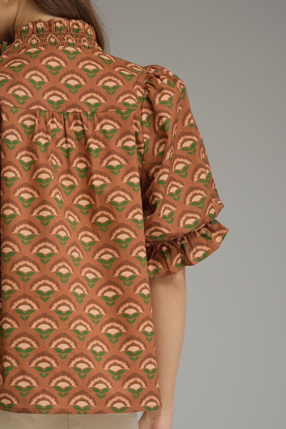 A close-up of a model wearing a short-sleeve blouse with a brown, green, and beige geometric fan pattern, featuring a smocked high collar and ruffled sleeves.