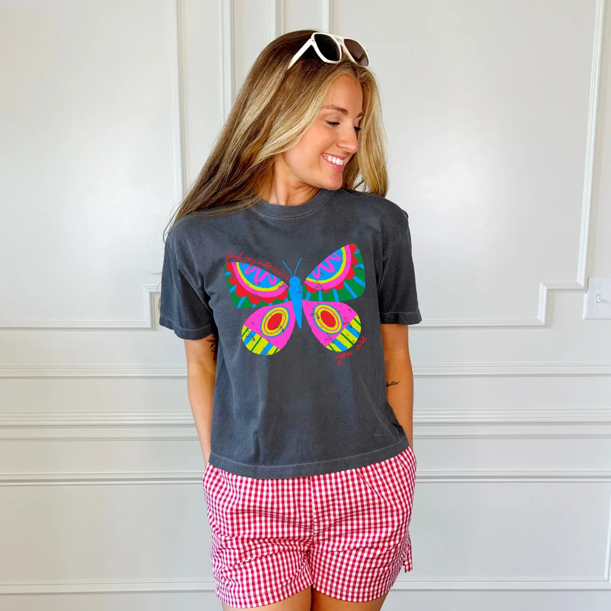 A woman wearing a dark gray graphic t-shirt with a colorful butterfly print and red-and-white gingham shorts, standing against a white paneled wall.