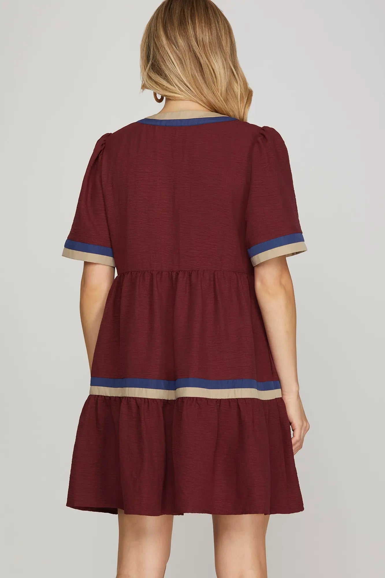 Back view of a burgundy short dress featuring tiered layers and contrast navy and taupe trim on the sleeves and hem, styled with loose waves.