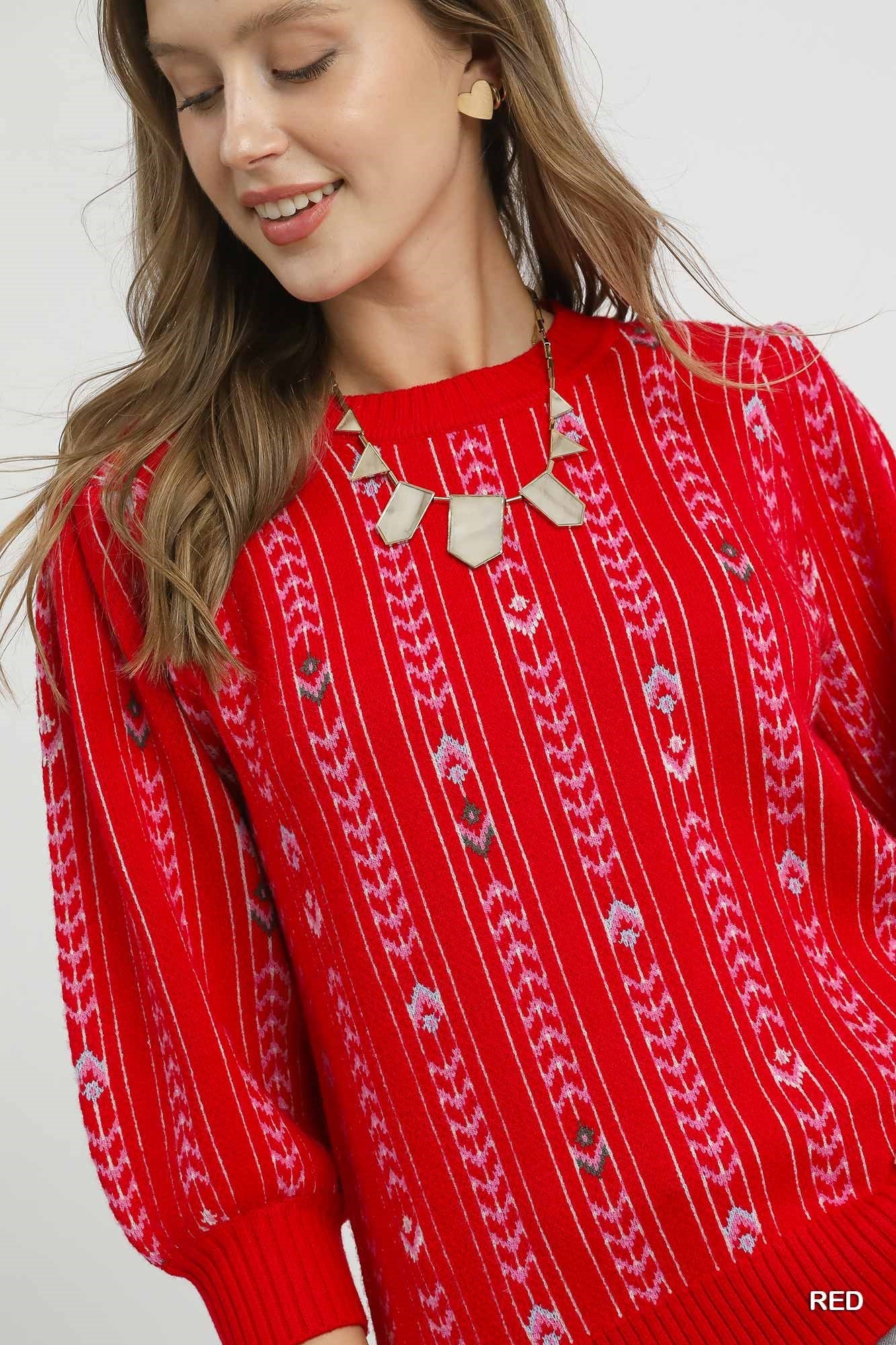 Woman wearing a red knit sweater with embroidered fair isle-inspired pattern, puff sleeves, and ribbed cuffs, styled with a statement necklace and heart-shaped earrings.
