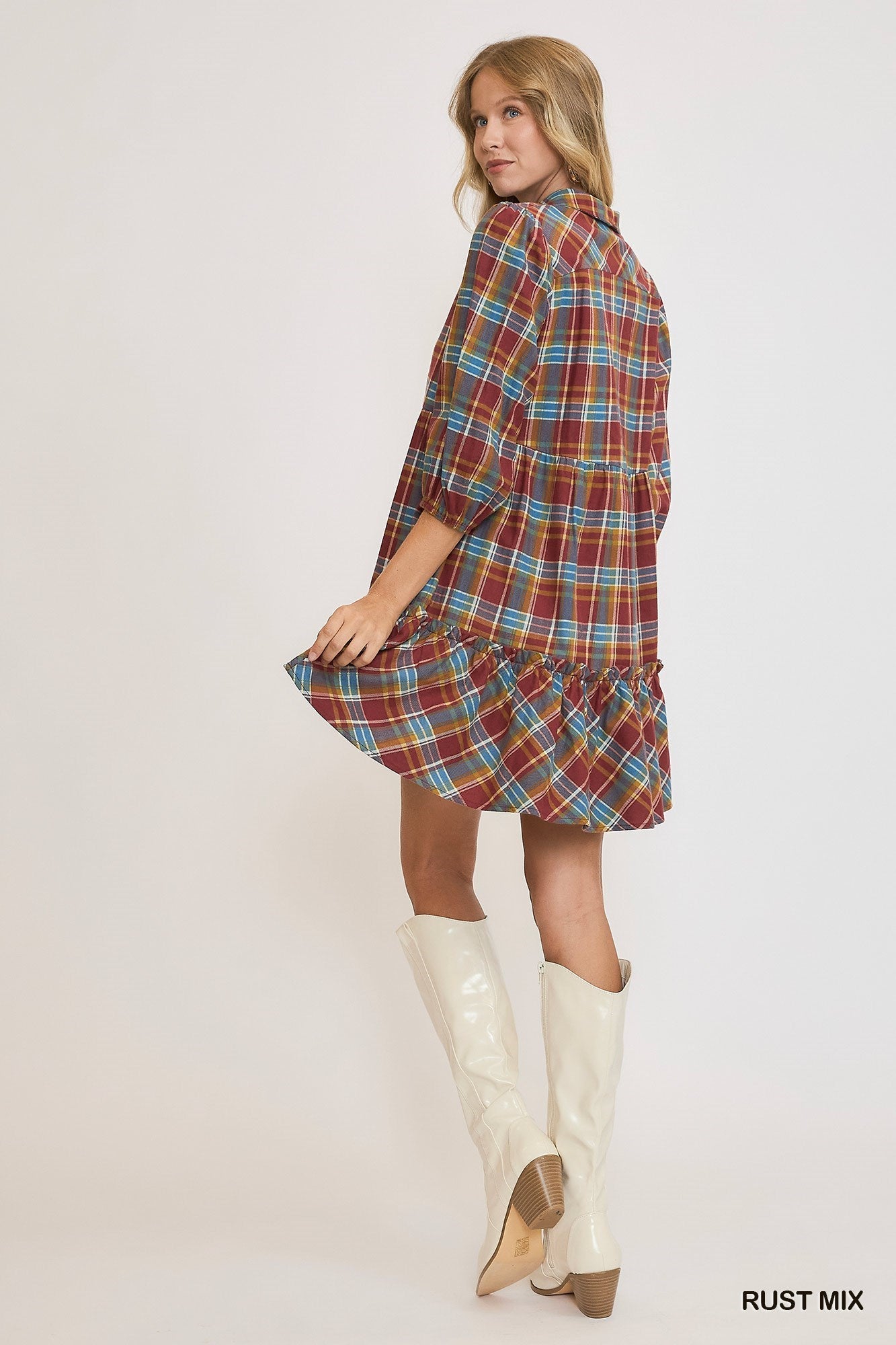 Model wearing a rust mix plaid mini dress with a ruffle hem, button neckline, and puff sleeves, styled with white knee-high boots against a light background.