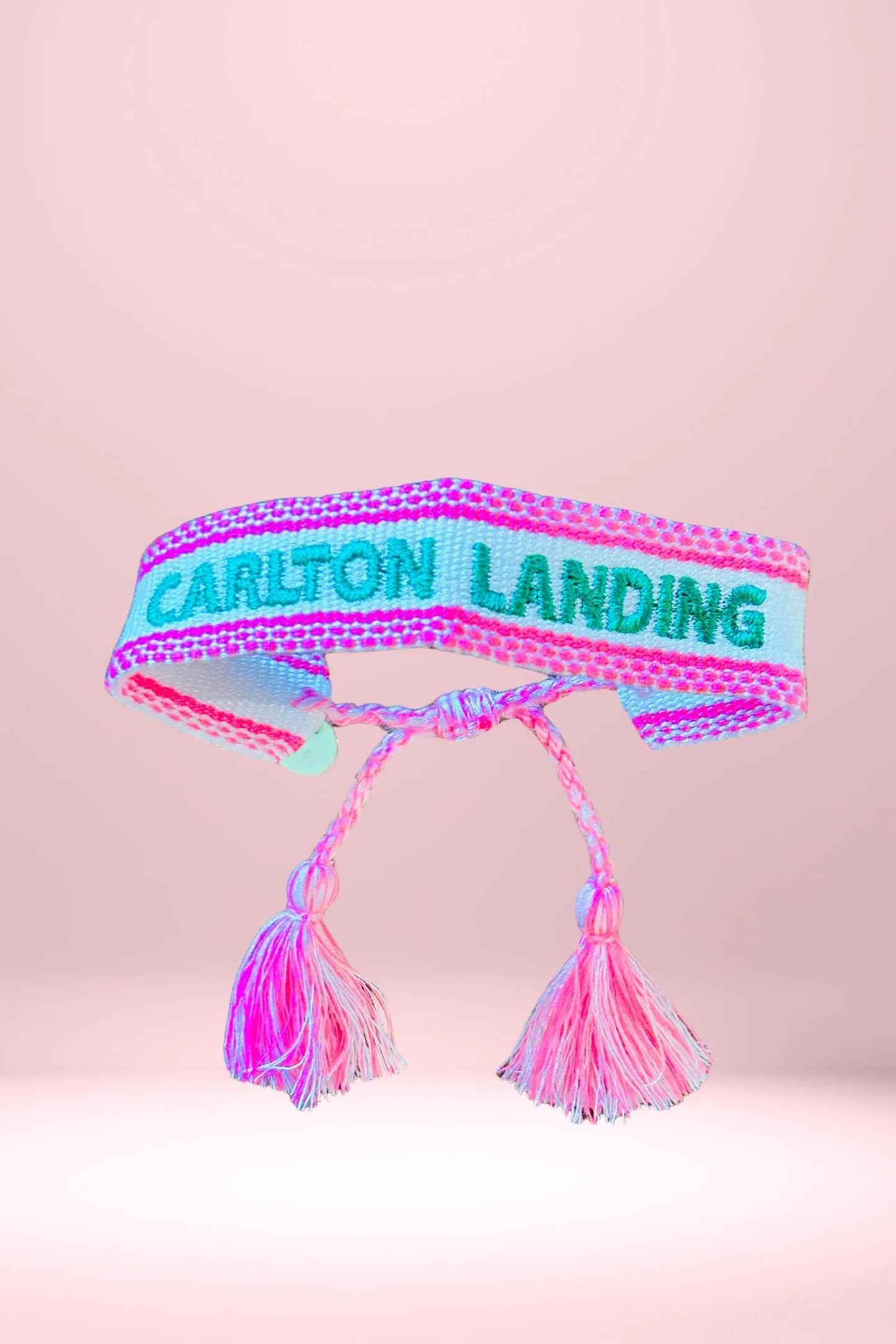 Pink and turquoise woven friendship bracelet with tassels and 'Carlton Landing' text on a pink background
