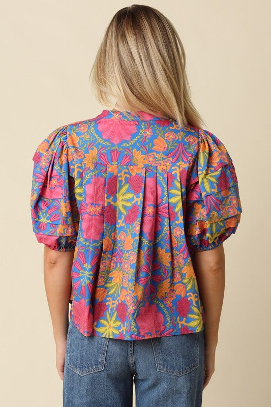 A woman viewed from the back wearing a colorful short-sleeve blouse with a vibrant floral and paisley print in pink, blue, orange, and yellow, paired with blue jeans against a solid beige background.
