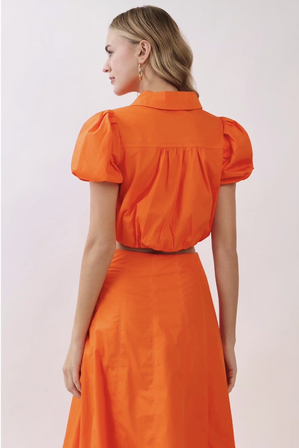 A woman shown from the back wearing a bright orange two-piece outfit consisting of a short puff-sleeve collared top and a matching high-waisted skirt, against a plain light background.
