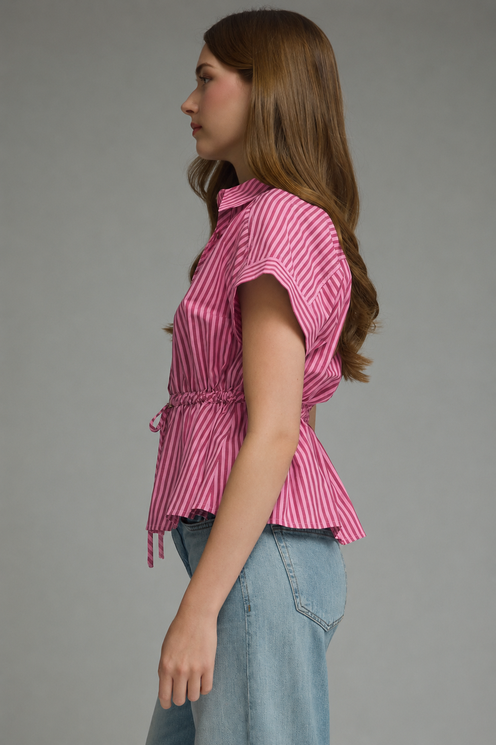 A woman shown in side profile wearing a pink and red vertically striped short-sleeve blouse with a drawstring waist, paired with light blue jeans, against a solid gray studio background.