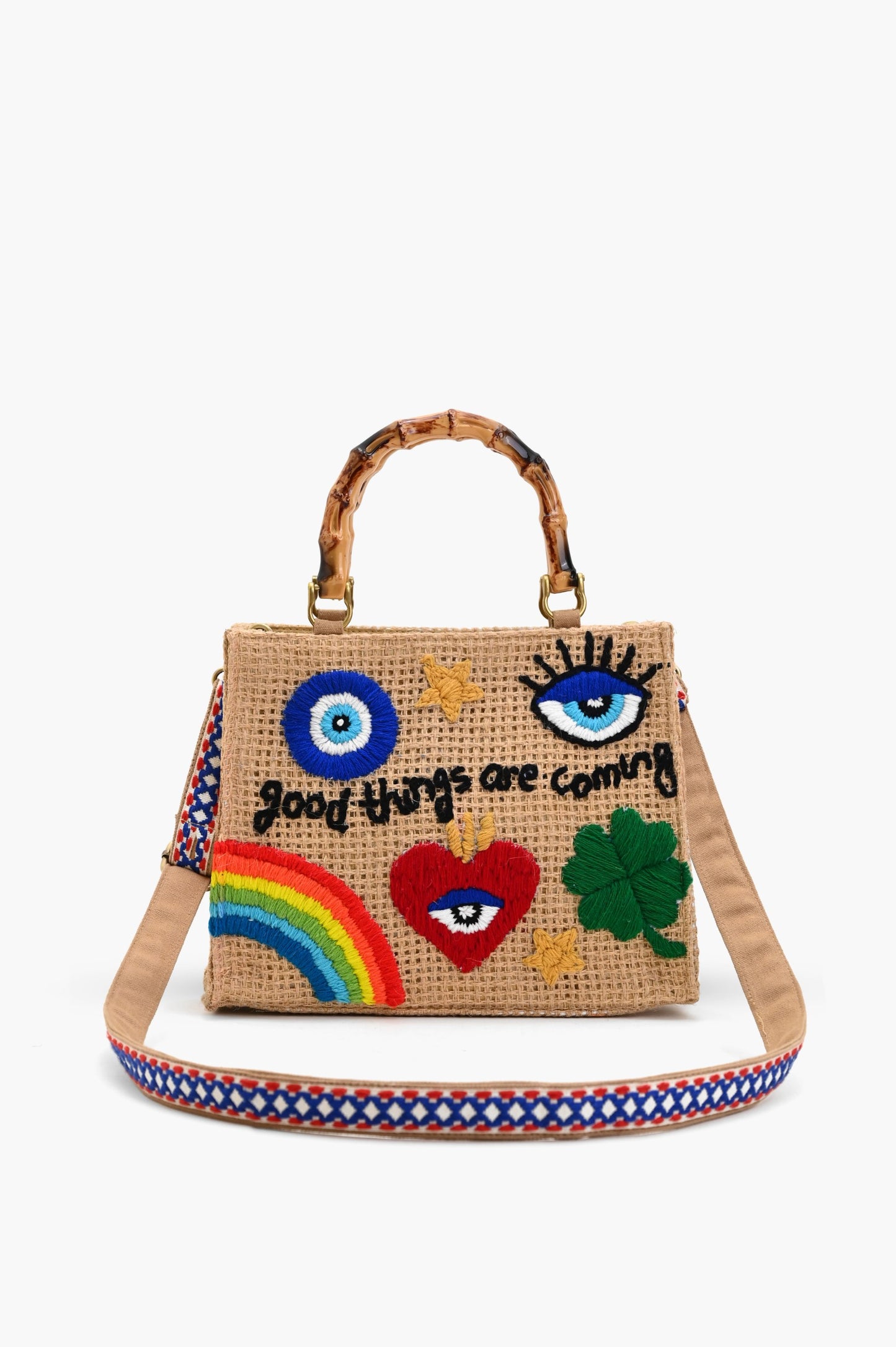 Natural woven tote bag with bamboo-style handles and colorful embroidered designs including a rainbow, heart, clover, eyes, and the phrase “good things are coming.”
