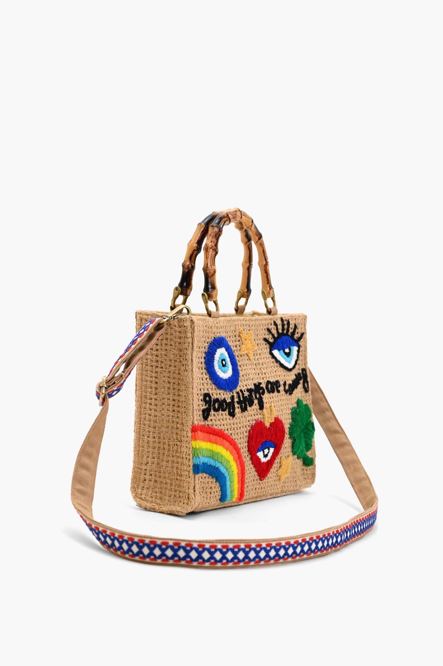 Natural woven tote bag with bamboo-style handles and colorful embroidered designs including a rainbow, heart, clover, eyes, and the phrase “good things are coming.”
