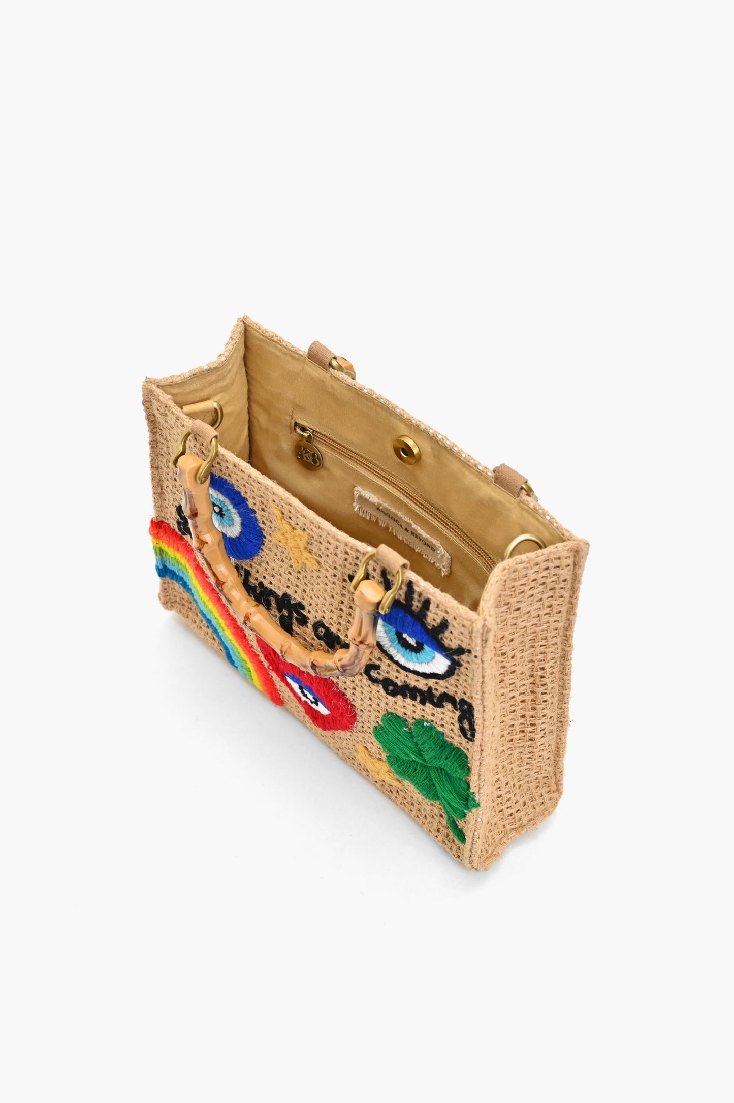 Natural woven tote bag with bamboo-style handles and colorful embroidered designs including a rainbow, heart, clover, eyes, and the phrase “good things are coming.”