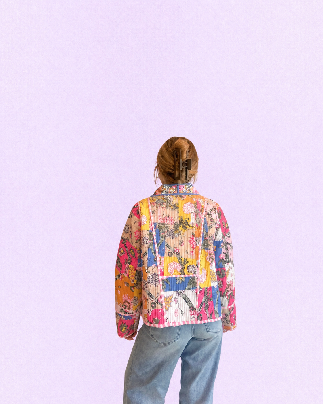 A person viewed from behind wearing a colorful patchwork-print jacket and light blue jeans against a solid light purple background.