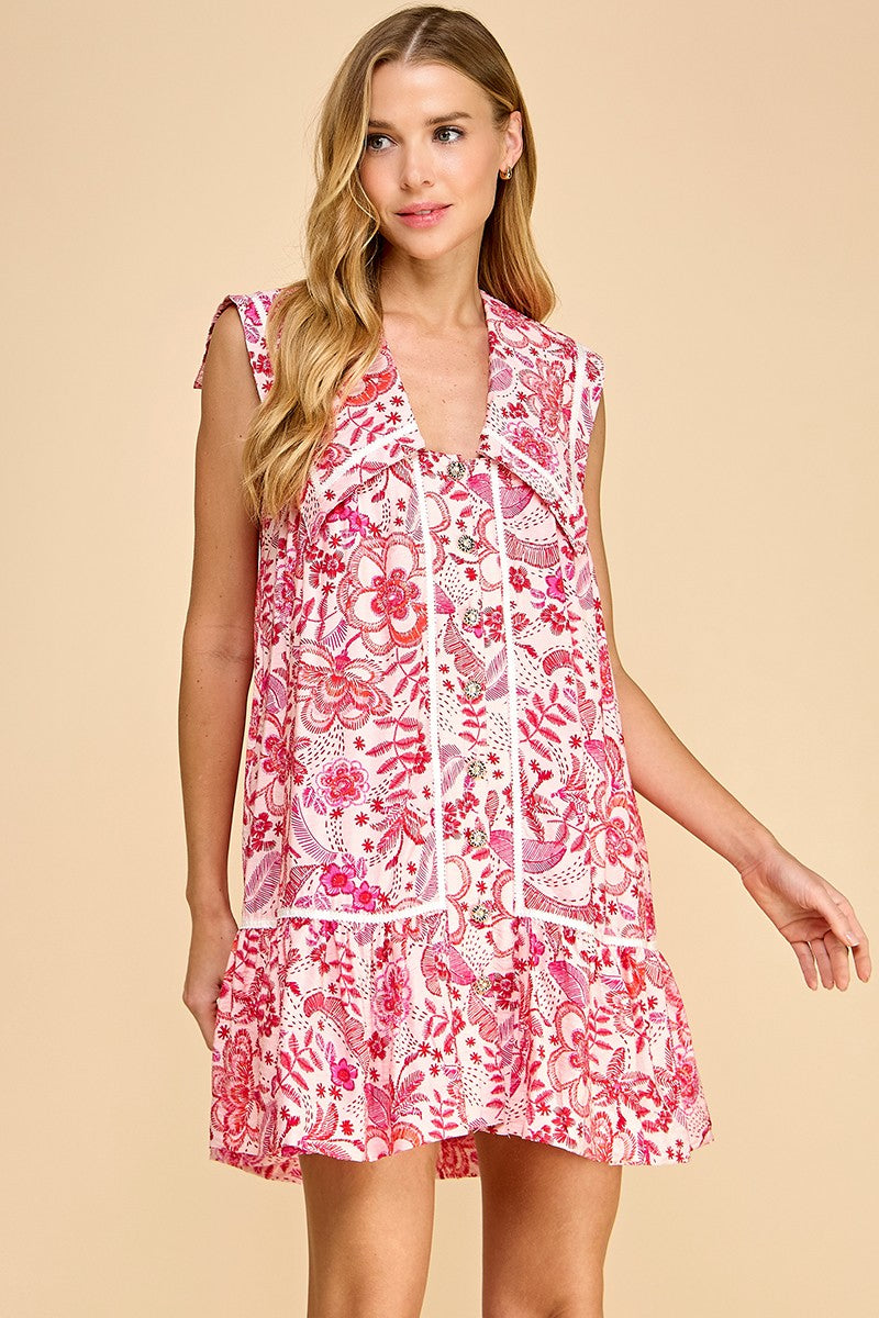 Woman wearing a pink and white floral print sleeveless mini dress with a tiered hem and oversized sailor collar, featuring front ties and side pockets.