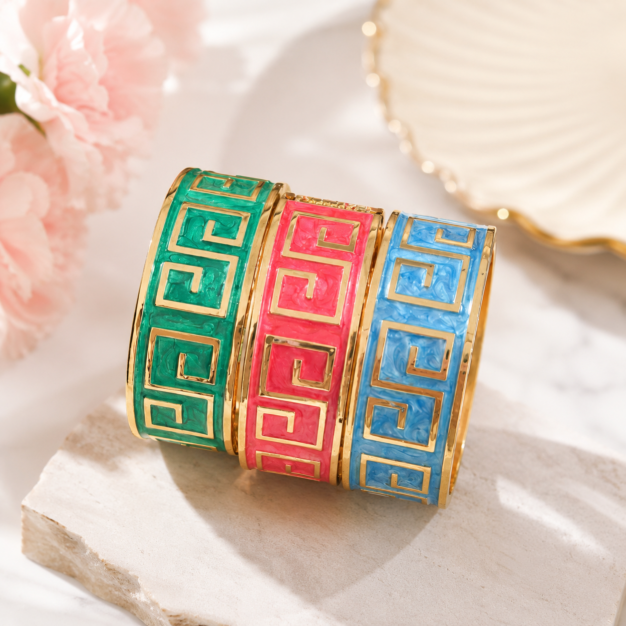 Three gold-plated cuff bracelets with Greek key patterns in green, pink, and blue enamel, arranged on a marble surface with soft pink flowers in the background.