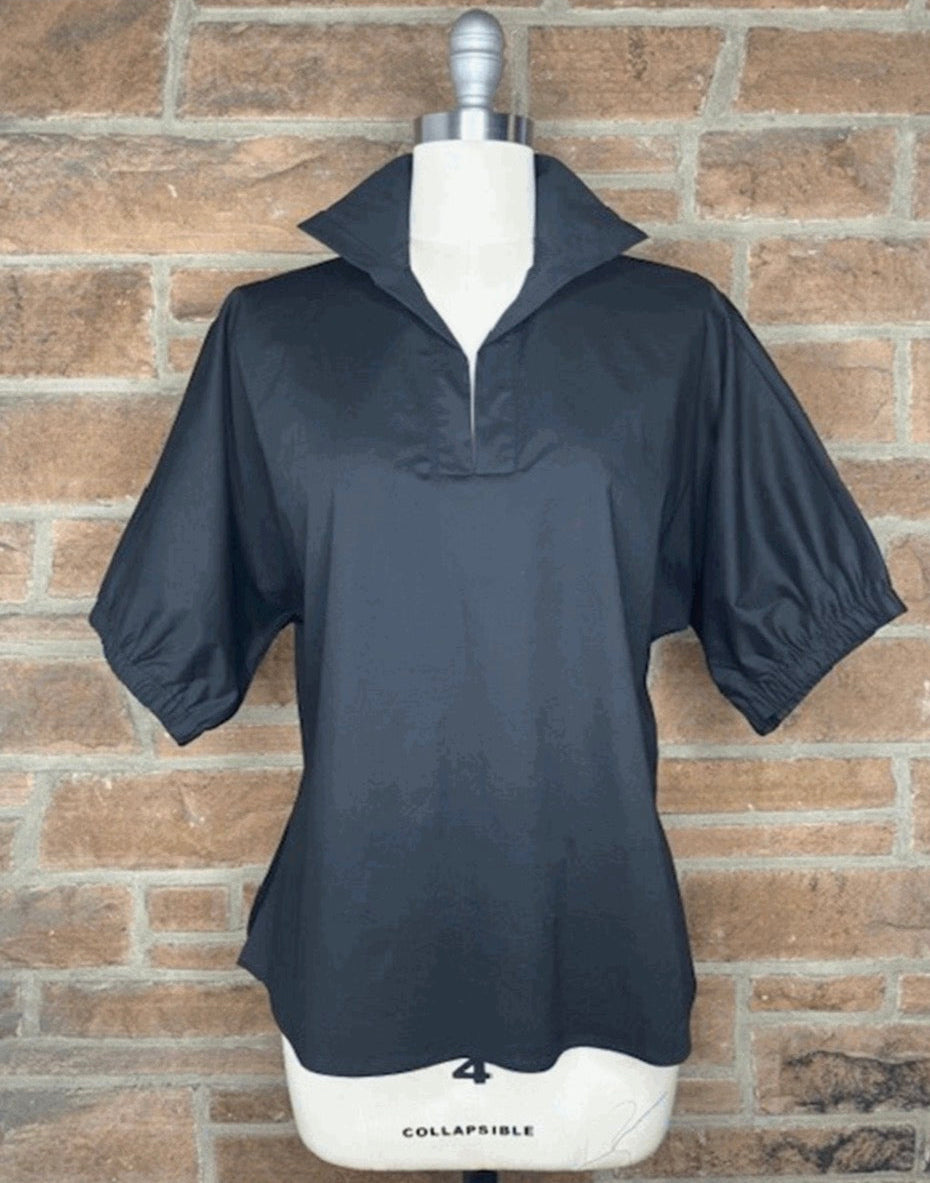 A black short-sleeve collared top displayed on a white mannequin torso against a brown brick wall. The top has a stand collar, a short front zipper, and gathered elastic cuffs.