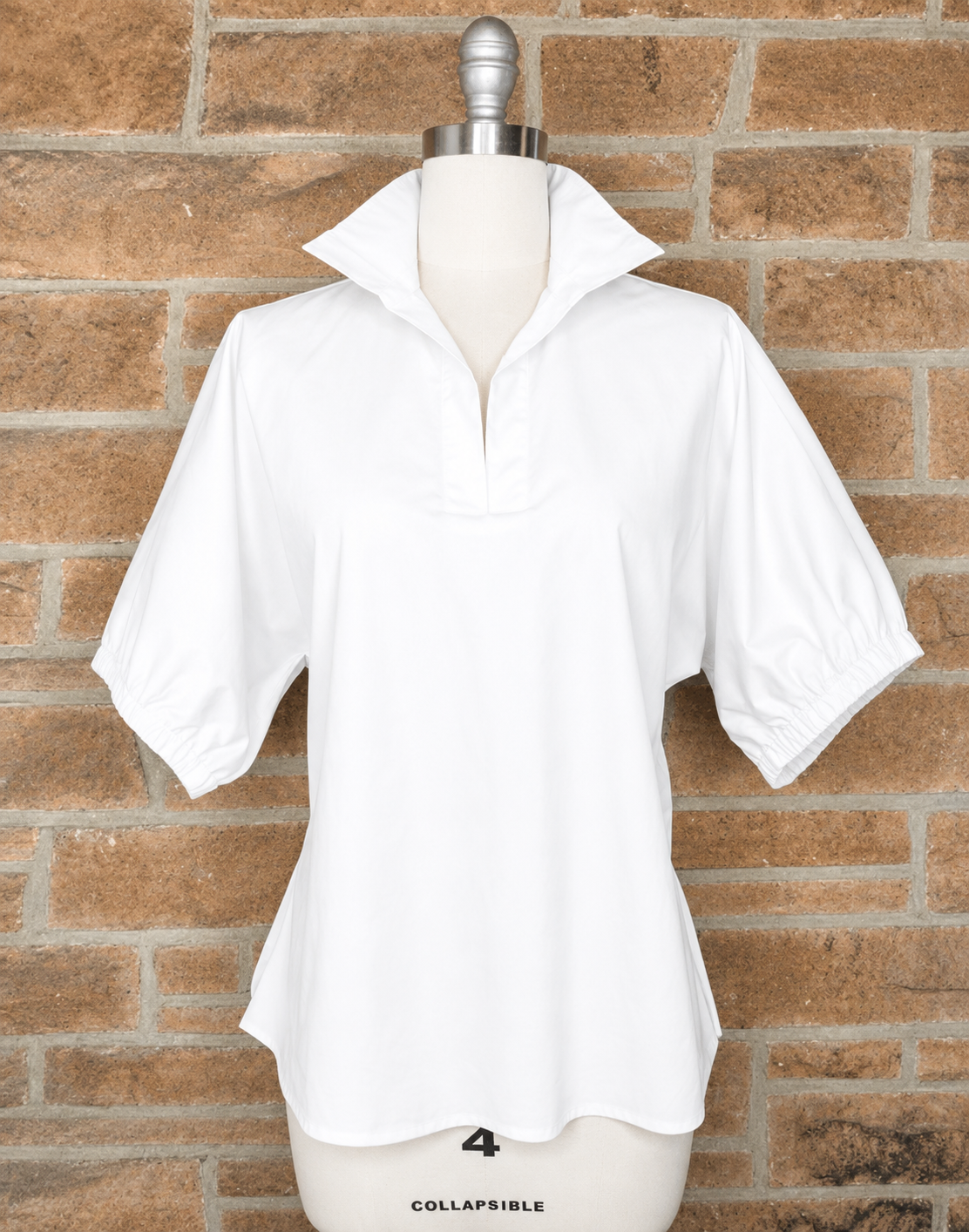 A white short-sleeve collared blouse with a partial zip and elasticized cuffs, displayed on a mannequin torso against a brown brick wall background.