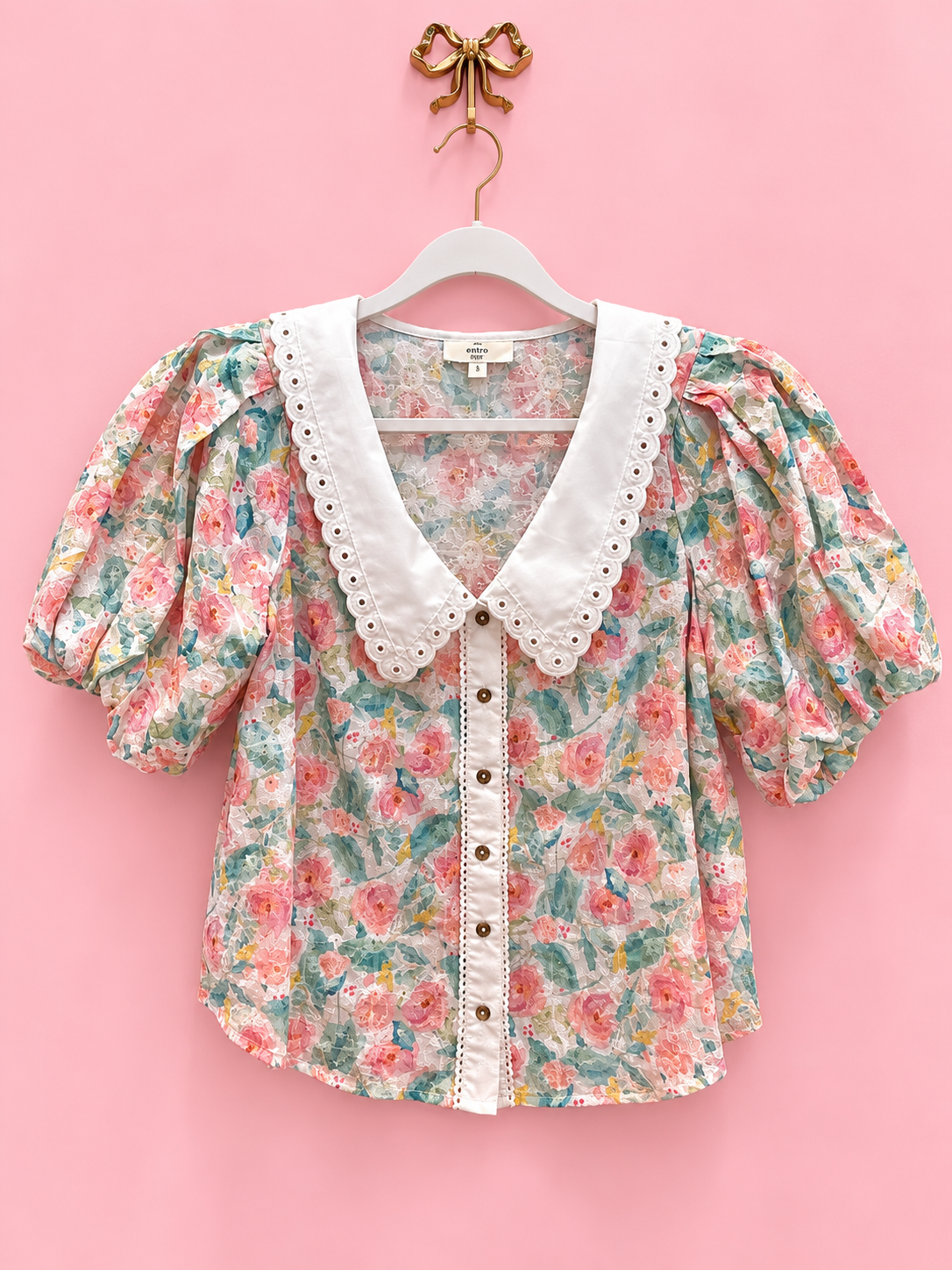 A short-sleeve floral blouse with puff sleeves and a white eyelet Peter Pan collar, hanging on a white hanger against a solid pink background.