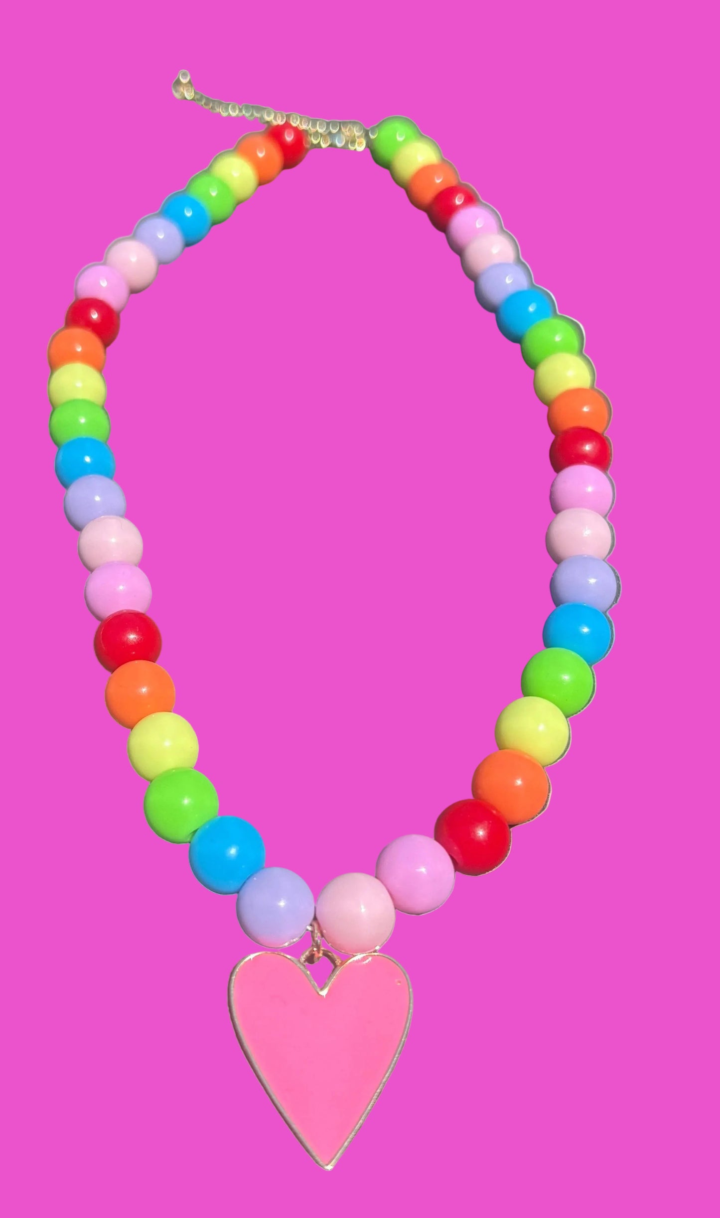 A colorful beaded necklace with rainbow-colored round beads and a pink heart-shaped pendant, displayed on a dark gray surface.