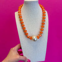 An orange beaded necklace with a central white pearl-like bead, displayed on a white mannequin bust against a bright pink background. A hand with pink nail polish is visible at the bottom left holding the display bust.