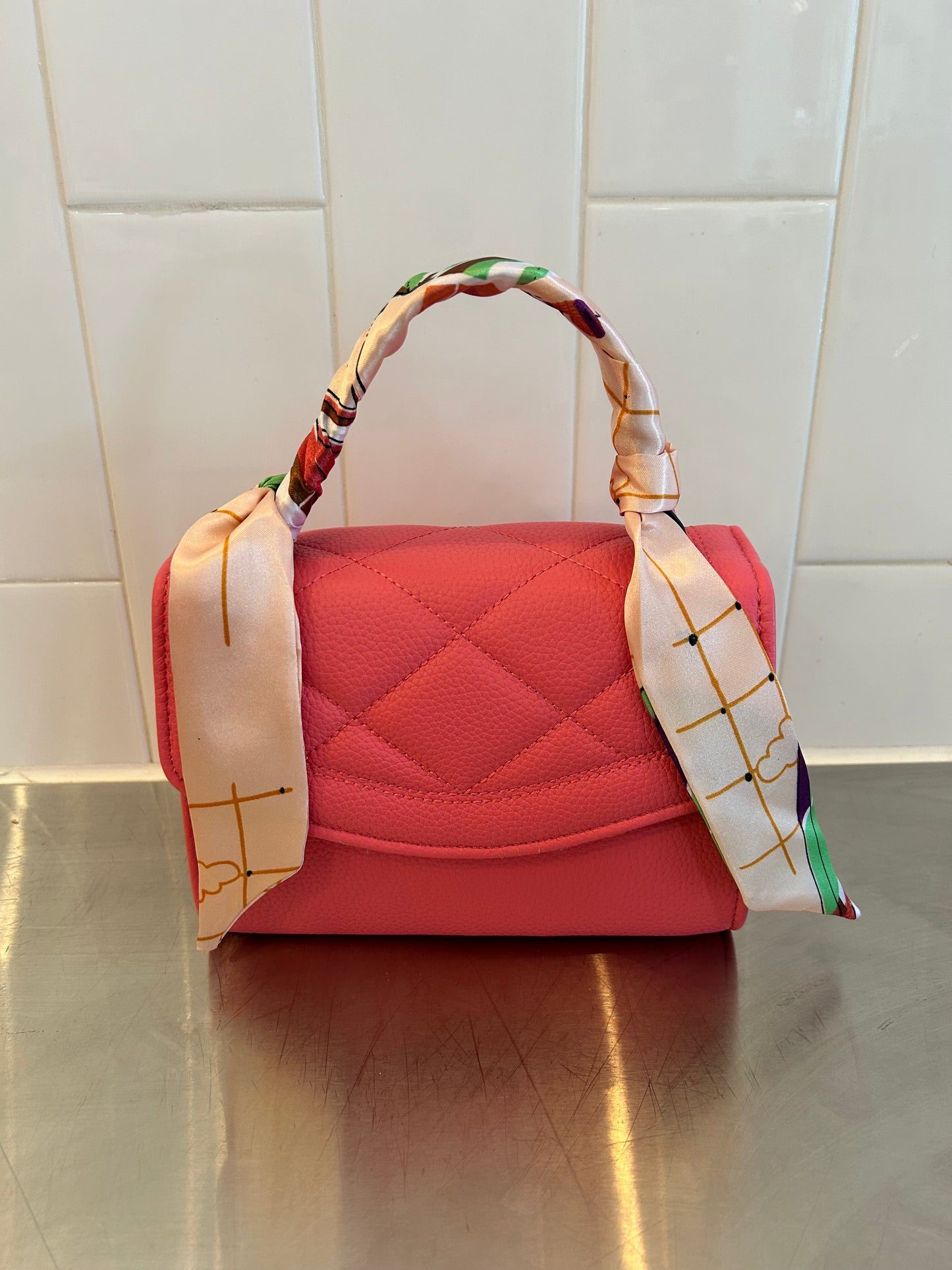 Pink quilted handbag with patterned scarf handle, on metal counter with white tile background