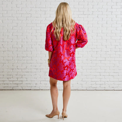 Woman in a pink floral puff sleeve mini dress and nude heels standing by white brick wall
