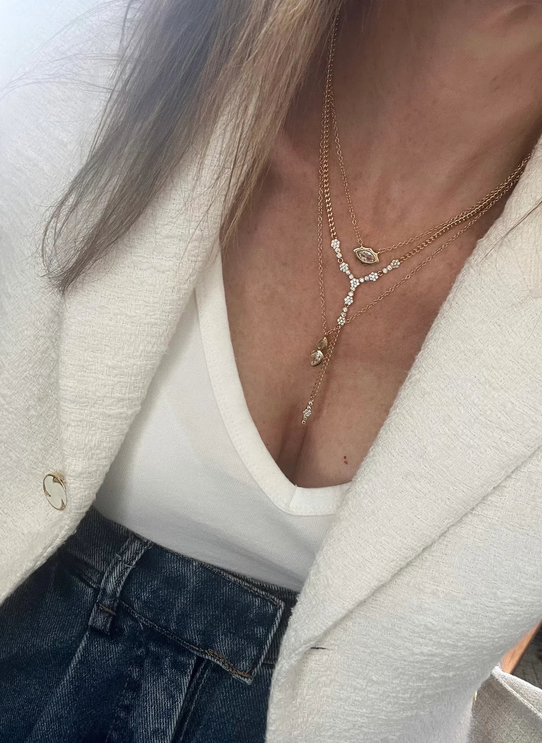 Layered gold necklaces with crystals worn over a white top, cream blazer, and denim jeans