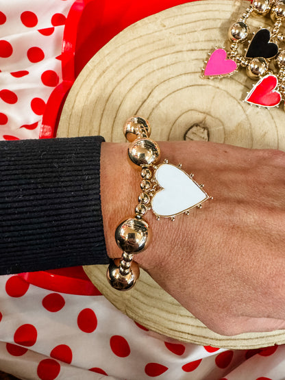 Gold beaded bracelet with white heart charm on wrist, polka dot background, trendy jewelry