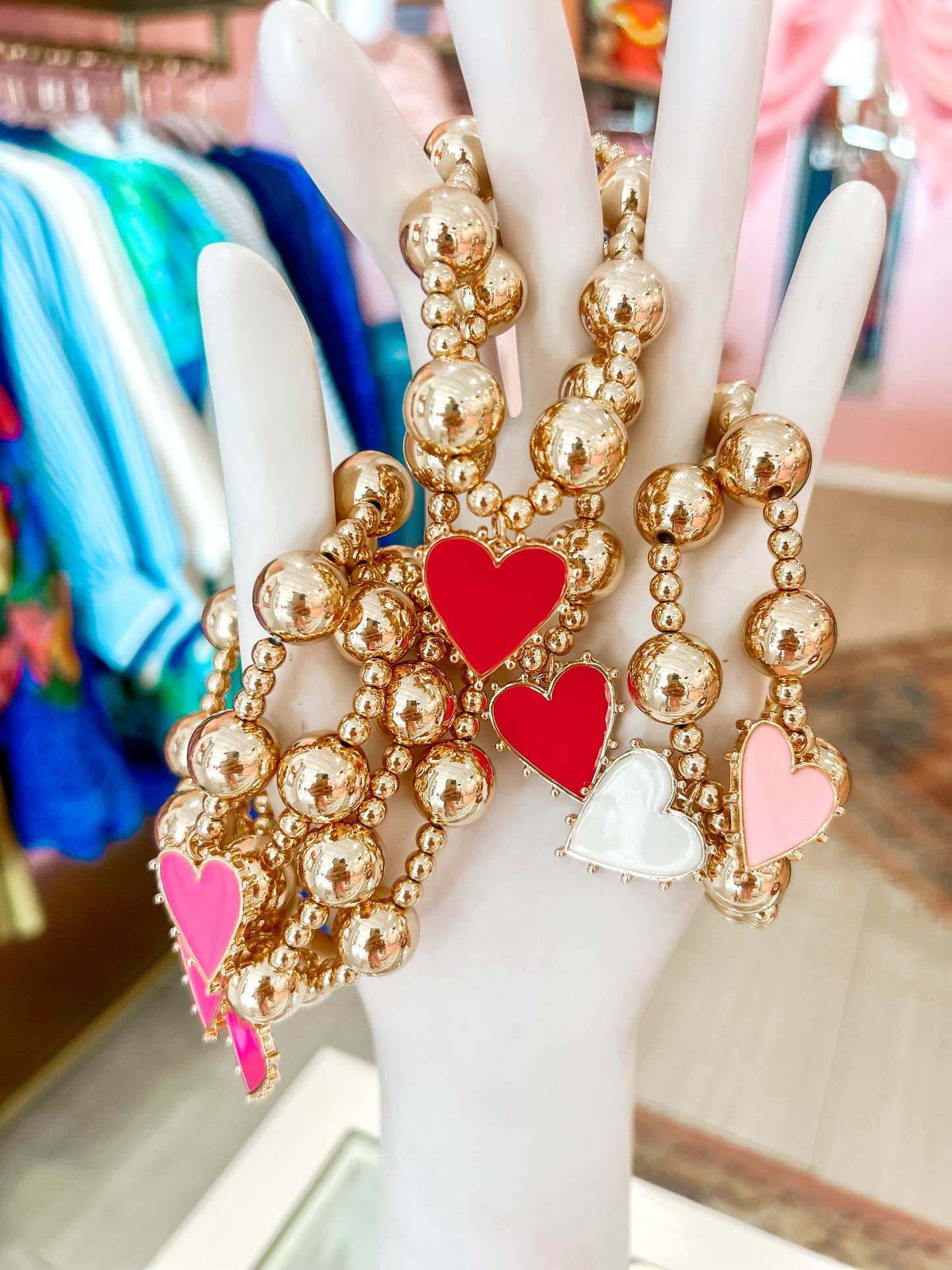 Gold beaded bracelets with red, pink, and white heart charms displayed on a hand mannequin
