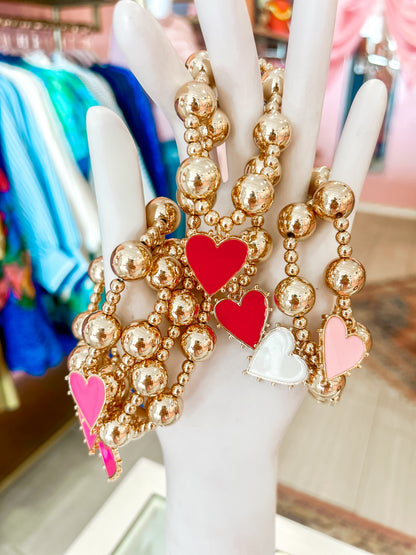 Gold beaded bracelets with red, pink, and white heart charms displayed on a hand mannequin