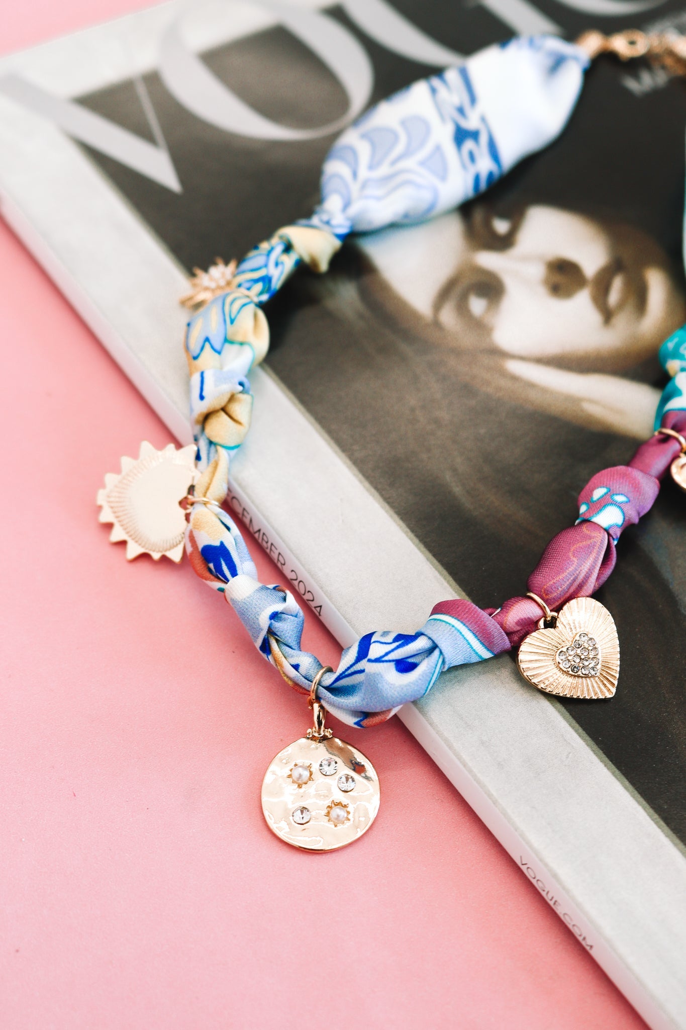 Colorful silk scarf necklace with gold charms, including heart and sun pendants, on magazine