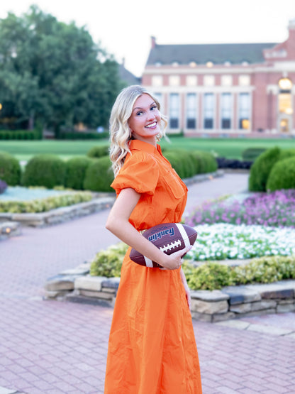 A woman posing outdoors in a bright orange dress while holding a brown football with 'Homerun' written on it. The background shows a landscaped garden with a brick building.