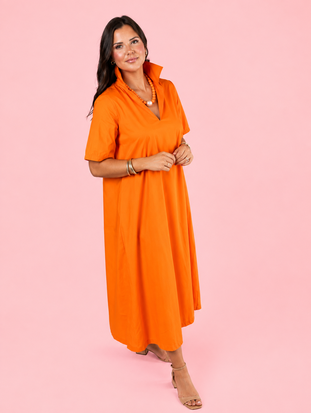 A woman modeling a bright orange short-sleeve midi dress with a split neckline and V-neckline detail, standing against a solid pink background.