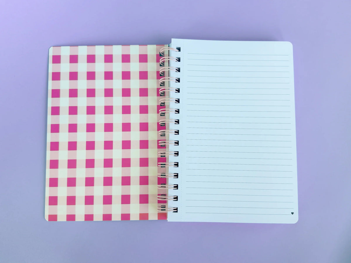 An open spiral-bound notebook with a pink and white gingham patterned cover on the left and lined blank pages on the right, photographed on a solid lavender background.