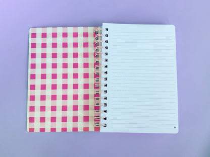 An open spiral-bound notebook with a pink and white gingham patterned cover on the left and lined blank pages on the right, photographed on a solid lavender background.