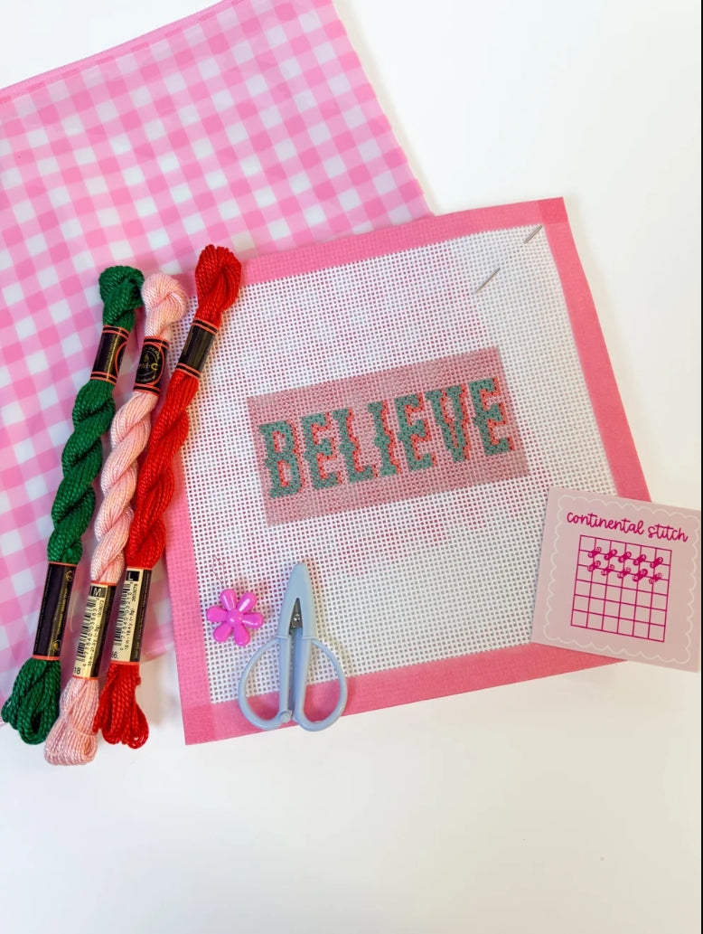 A flat lay of embroidery supplies on a white surface, including three skeins of thread in green, light pink, and red, a pink-and-white gingham fabric piece, a white cross-stitch canvas with the word "BELIEVE" stitched in green, a pair of light blue scissors, a small pink flower, and a small instruction card.