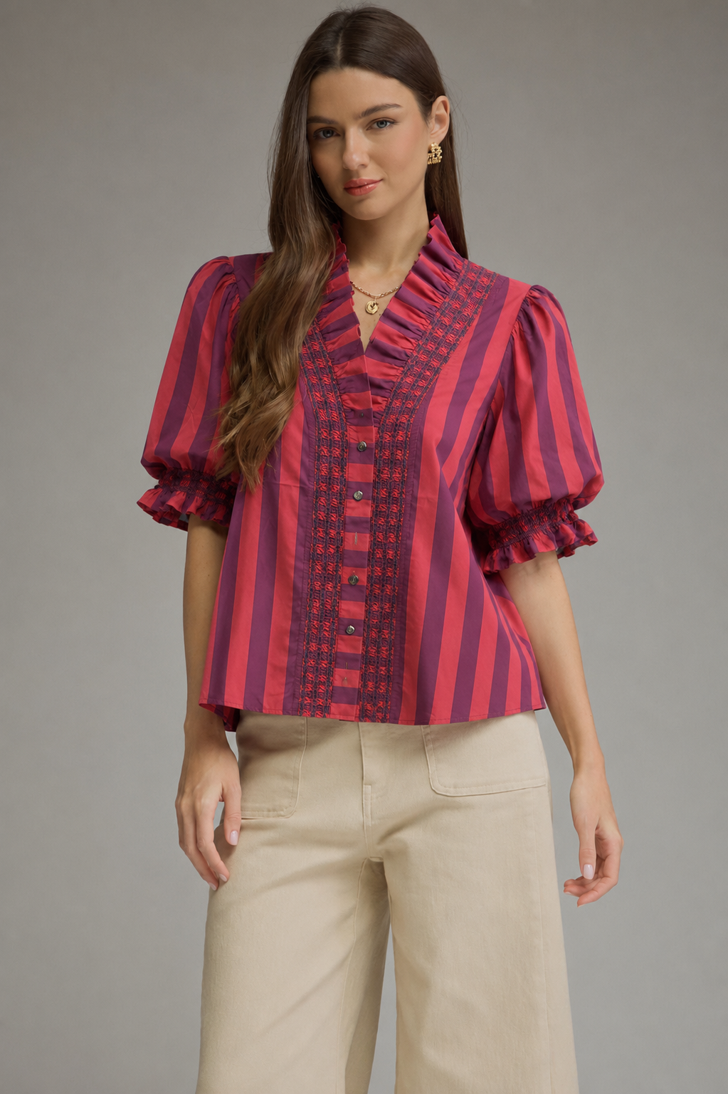 A woman modeling a short-sleeve button-front blouse with vertical pink and dark red stripes and puff sleeves, paired with light beige pants, against a plain gray studio background.
