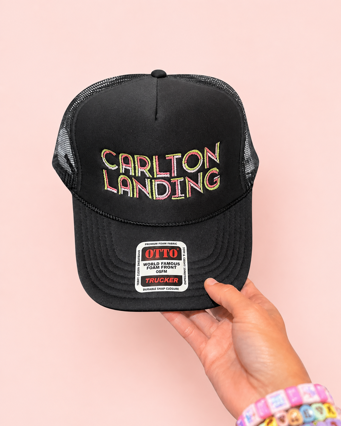 A black trucker hat with 'CARLTON LANDING' embroidered in a multicolor outline font on the front, held by a hand against a solid light pink background. The hat features a white 'OTTO' sticker on the brim.