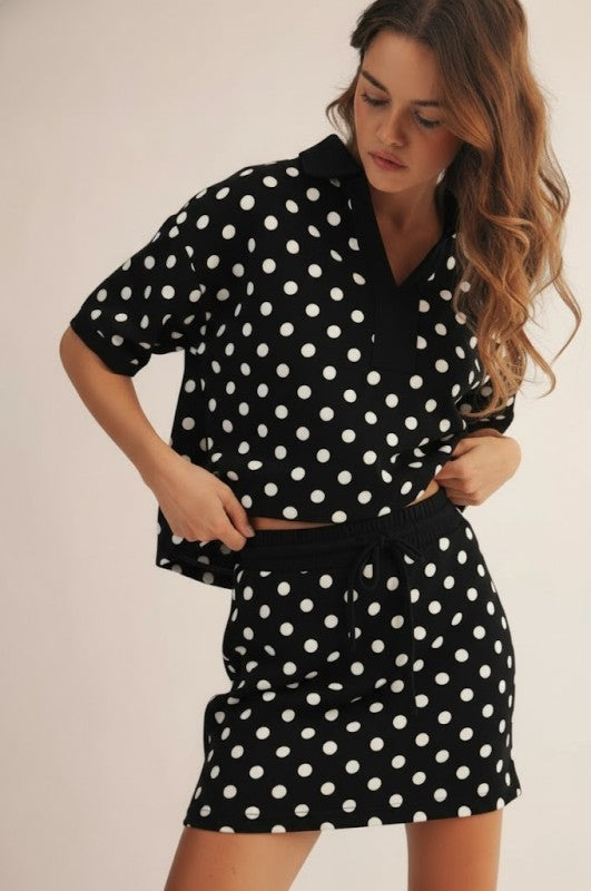 A woman modeling a matching black and white polka dot lounge set consisting of a short-sleeve collared top and a high-waisted mini skirt, against a plain light beige background.