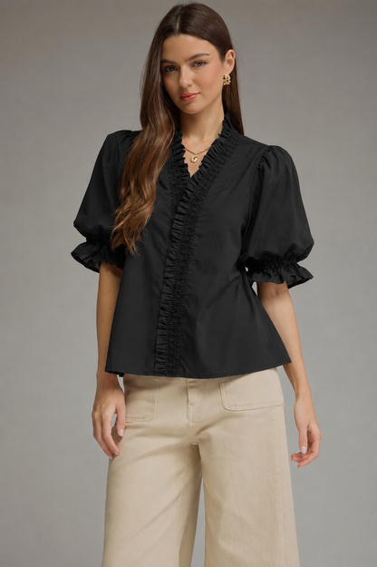 A woman modeling a black short-sleeve blouse with ruffled trim and puffed sleeves, paired with beige wide-leg pants, against a plain gray studio background.