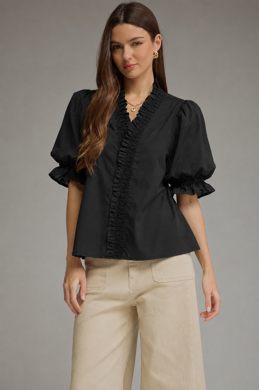 A woman modeling a black short-sleeve blouse with ruffled trim and puffed sleeves, paired with beige wide-leg pants, against a plain gray studio background.