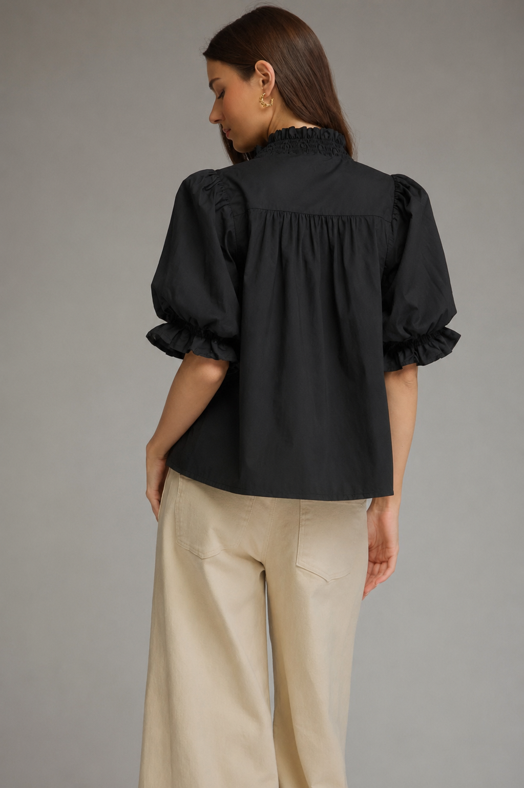 A woman shown from the back wearing a black puff-sleeve blouse with ruffled trim and beige wide-leg trousers, photographed against a plain gray studio background.