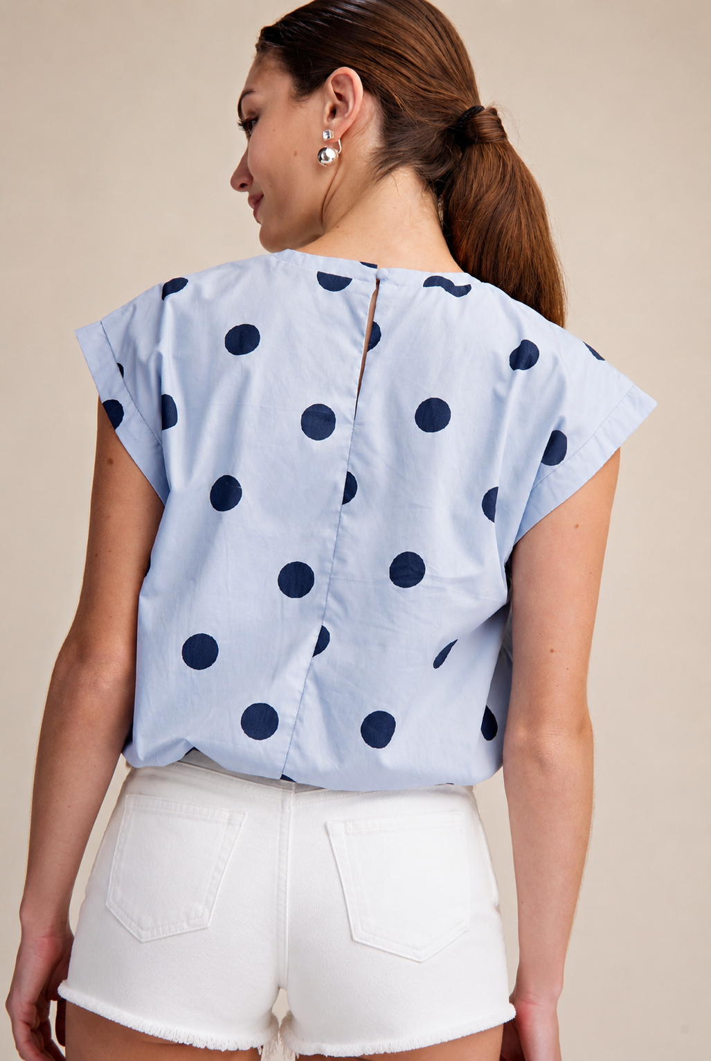 Woman wearing a light blue blouse with dark polka dots and white shorts on a beige background