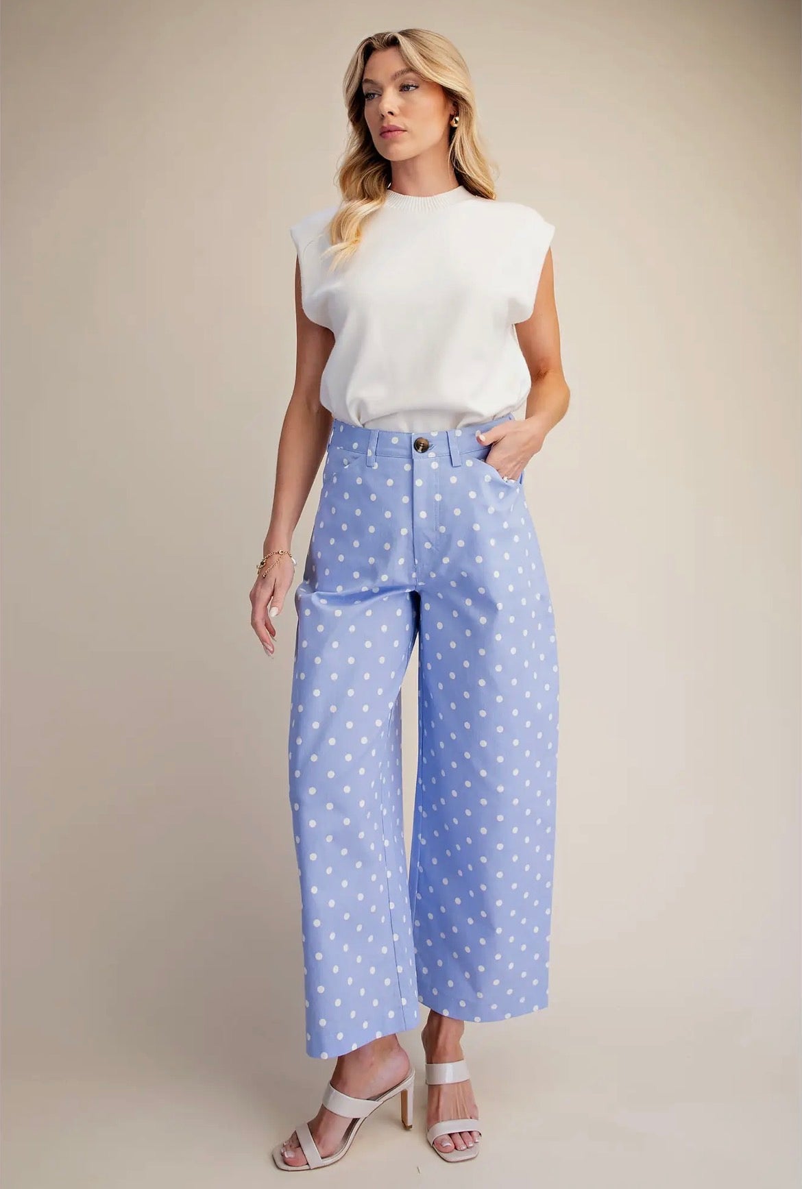A woman modeling a white sleeveless top and light blue wide-leg cropped pants with white polka dots, paired with white heeled sandals, against a solid beige studio background.
