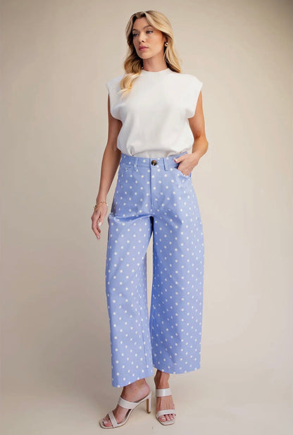 A woman modeling a white sleeveless top and light blue wide-leg cropped pants with white polka dots, paired with white heeled sandals, against a solid beige studio background.