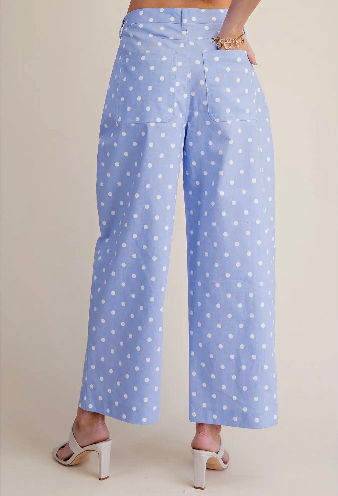 A person shown from the back wearing light blue cropped pants with a white polka dot pattern, paired with white heeled sandals against a plain beige studio background.