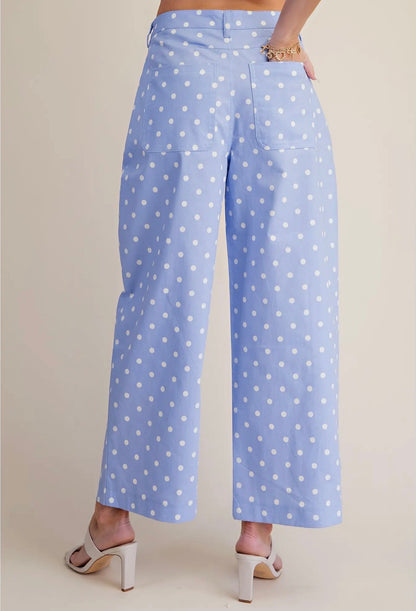 A person shown from the back wearing light blue cropped pants with a white polka dot pattern, paired with white heeled sandals against a plain beige studio background.