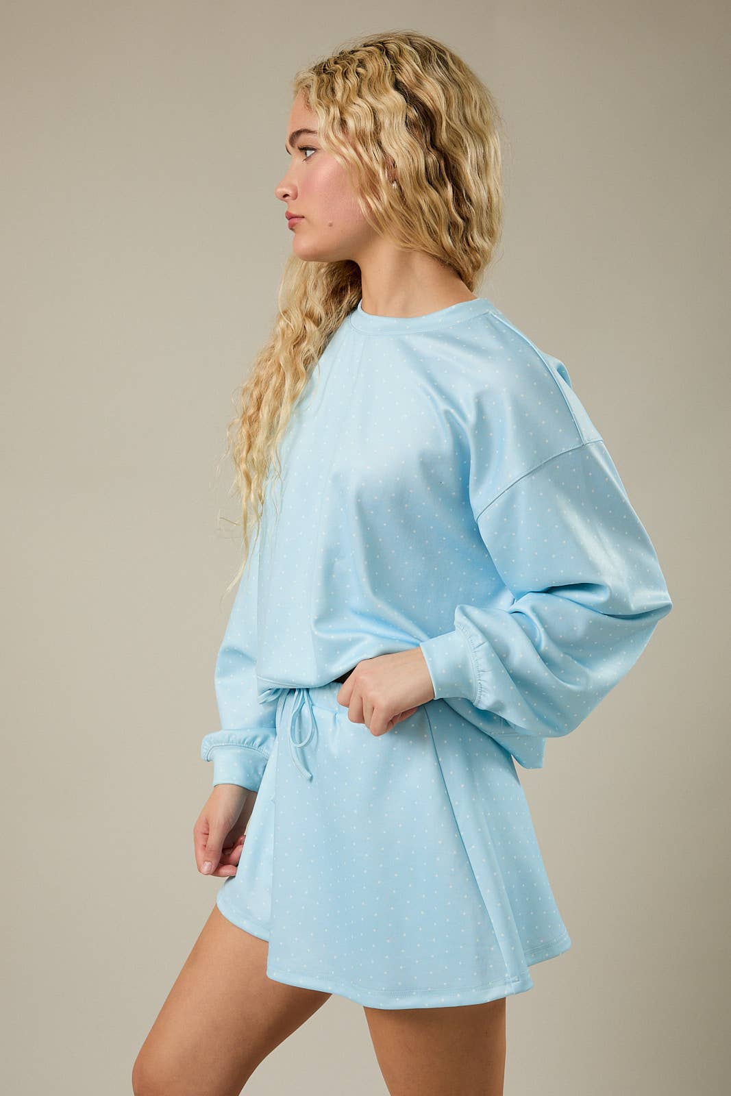 A woman with long blonde wavy hair is shown in profile wearing a light blue oversized sweatshirt and matching shorts set against a plain beige studio background.
