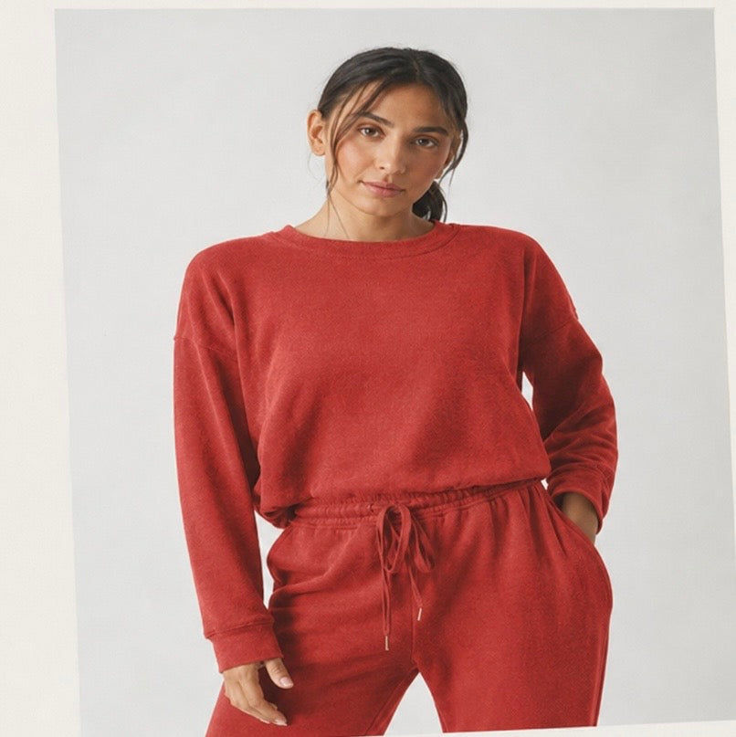 A woman wearing a matching rust-red sweatshirt and jogger pants set, photographed against a plain light gray studio background.