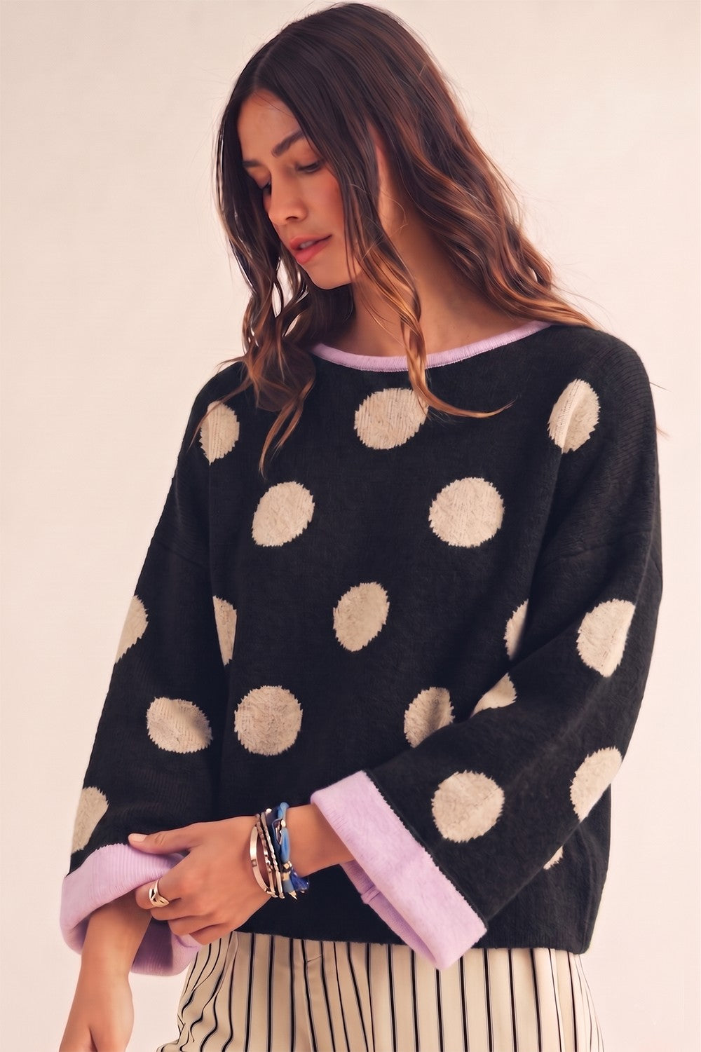 A woman wearing a black knit sweater with large beige polka dots and light purple cuffs, paired with striped pants, against a plain beige background.