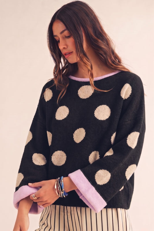 A woman wearing a black knit sweater with large beige polka dots and light purple cuffs, paired with striped pants, against a plain beige background.