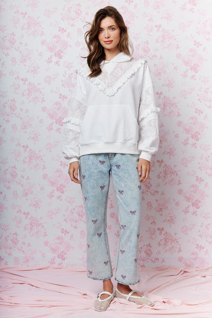 A woman stands against a pink floral backdrop wearing a white oversized hoodie with lace ruffle details, light-wash blue jeans with small butterfly prints, and beige slip-on shoes.