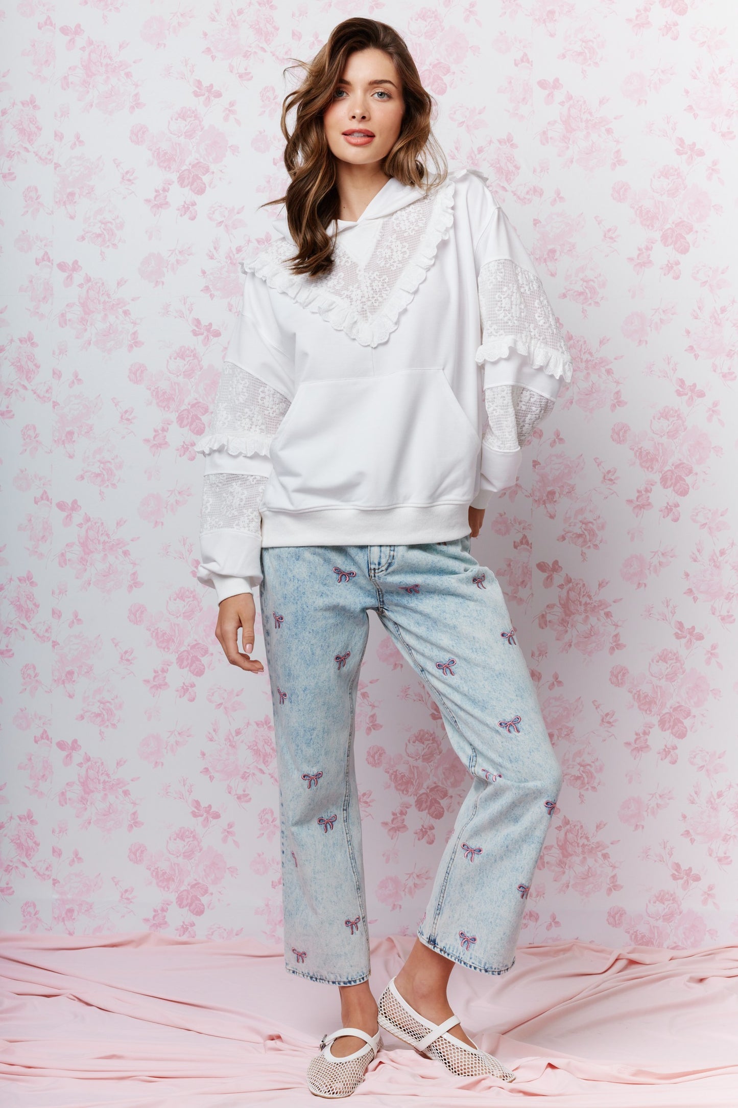 A woman modeling a white ruffled long-sleeve hoodie and light-wash blue jeans with butterfly prints, standing against a pink floral backdrop.
